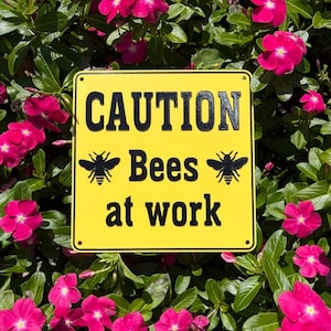 Può includere: Un cartello quadrato giallo con testo nero che recita CAUTION Bees at work, caratterizzato da una grafica di un'ape nera su ciascun lato della parola Bees. Il cartello ha piccoli fori circolari in ogni angolo per il montaggio e una superficie liscia.