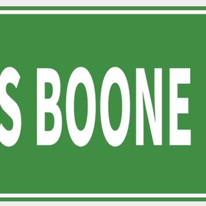 Pode incluir: Uma placa de rua verde com o texto branco "USS BOONE DR". A placa tem uma borda branca e é montada com dois rebites. A placa é retangular.