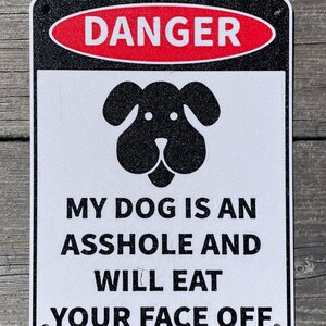 Op de afbeelding: Een wit bord met zwarte tekst en een zwart silhouet van een hondenkop. Het bord staat "DANGER" in een rode ovaal bovenaan en "MY DOG IS AN ASSHOLE AND WILL EAT YOUR FACE OFF" in hoofdletters.