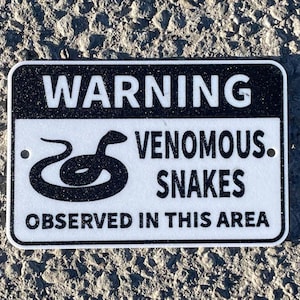 Peut inclure: Un panneau d'avertissement noir et blanc avec le texte "WARNING VENOMOUS SNAKES OBSERVED IN THIS AREA". Une silhouette noire d'un serpent se trouve au centre du panneau.