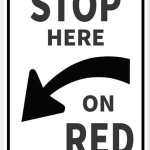 May include: A rectangular white traffic sign with black text and graphics. The sign reads "STOP HERE ON RED" with a curved arrow pointing left. The sign has a black border and two rivets.
