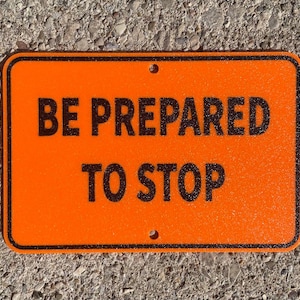 May include: Orange and black sign with the text "BE PREPARED TO STOP" on a concrete surface.