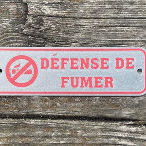 May include: A silver sign with red text and a red no smoking symbol. The sign reads "Défense de Fumer".
