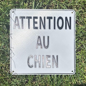 以下が含まれることがあります： 「ATTENTION AU CHIEN」の文字がエンボス加工された、四角い銀色の金属製の標識。標識には丸みを帯びた角と、取り付け用の小さな穴があります。標識は緑の草の上に置かれています。