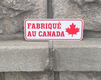 May include: A rectangular white sticker with a red border and text that reads "FABRIQUÉ AU CANADA" in red. A red maple leaf is on the right side of the sticker. The sticker is placed on a grey stone surface.