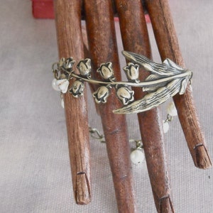 Puede incluir: Un brazalete plateado con un dise&ntilde;o de lirios del valle y cuentas blancas. El brazalete se muestra en una mano de madera.