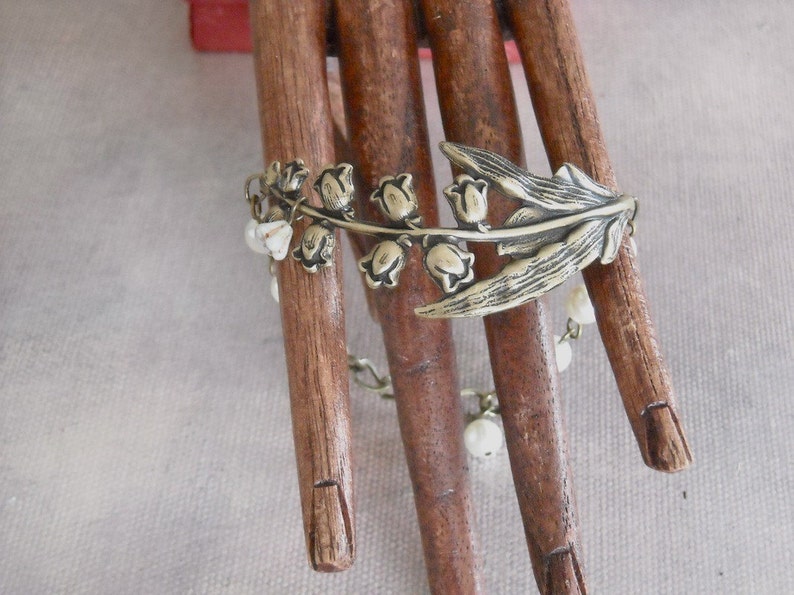 Puede incluir: Un brazalete plateado con un dise&ntilde;o de lirios del valle y cuentas blancas. El brazalete se muestra en una mano de madera.