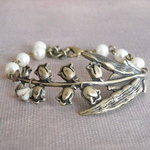 Puede incluir: Un delicado brazalete con un dije de bronce en forma de lirio de los valles con detalles de perlas blancas. El dije est&aacute; decorado con intrincados dise&ntilde;os florales y un acabado texturizado.