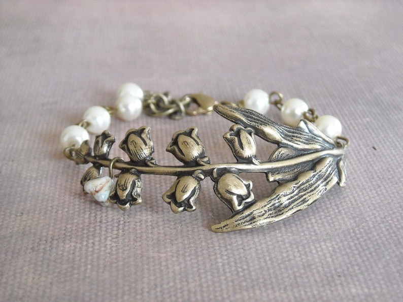 Puede incluir: Un delicado brazalete con un dije de bronce en forma de lirio de los valles con detalles de perlas blancas. El dije est&aacute; decorado con intrincados dise&ntilde;os florales y un acabado texturizado.