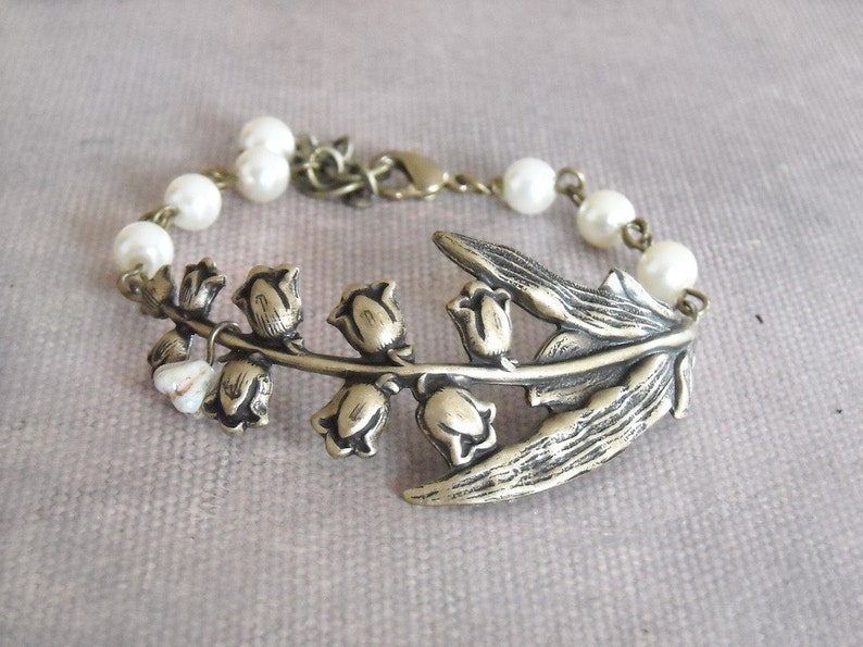 Puede incluir: Un delicado brazalete con un dije de lirio de los valles plateado y cuentas de perlas blancas.