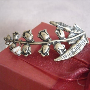 Puede incluir: Un brazalete de metal plateado con un dise&ntilde;o de flores de lirio de los valles. El brazalete est&aacute; sobre una caja de terciopelo rojo.