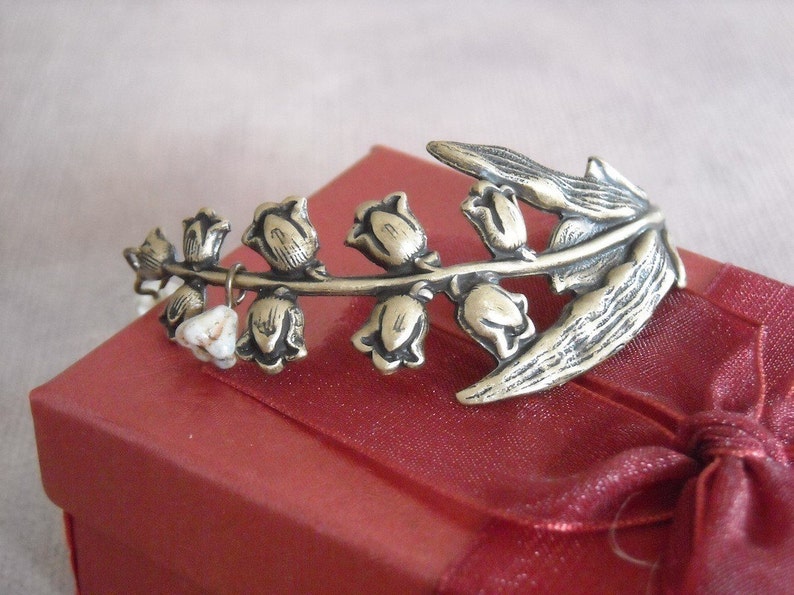 Puede incluir: Un brazalete de metal plateado con un dise&ntilde;o de flores de lirio de los valles. El brazalete est&aacute; sobre una caja de terciopelo rojo.