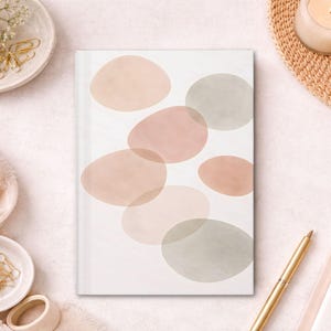 Puede incluir: Un diario blanco con un diseño abstracto de formas ovaladas superpuestas en tonos rosa y gris. El diario está rodeado de elementos decorativos, como una vela, un bolígrafo y pequeños cuencos con clips dorados.