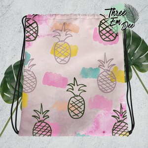 May include: A pink drawstring bag with a repeating pattern of black and white pineapple illustrations on a pink background with watercolor-like pink, blue, and yellow accents. The bag has black drawstrings.