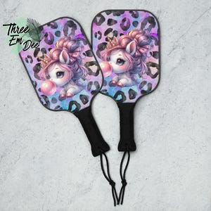 Puede incluir: Dos palas de pickleball con mango negro y diseño de unicornio. La cara de la pala tiene un fondo de estampado de leopardo en tonos morados y azules. El unicornio tiene pelo rosa y está soplando una burbuja.