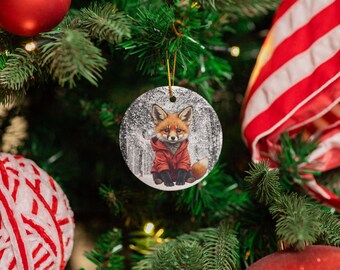 Peut inclure: Ornement de Noël rond représentant un renard en manteau rouge assis dans une forêt enneigée. L'ornement est suspendu par une ficelle dorée et se détache sur un fond de sapin de Noël vert, de boules rouges et d'un ruban rayé rouge et blanc.