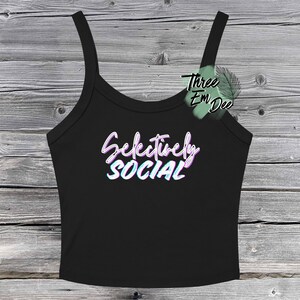 May include: Black tank top with thin straps. The words "Selectively SOCIAL" are written in a stylized, colorful font across the front. The background is a weathered, gray-toned wooden surface.