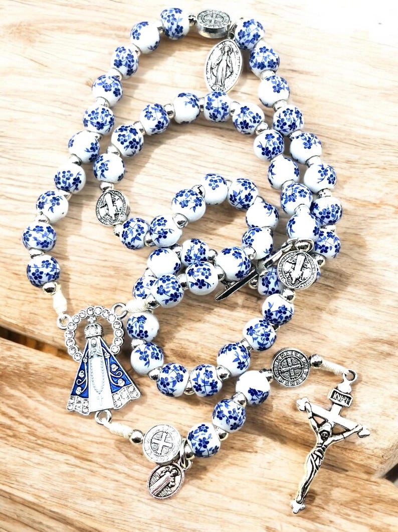 Puede incluir: Un rosario con cuentas blancas y azules con motivos florales. El rosario presenta detalles met&aacute;licos plateados, incluyendo un crucifijo, un colgante de la Virgen Mar&iacute;a y varios medallones religiosos. Las cuentas est&aacute;n dispuestas en un dise&ntilde;o tradicional de rosario.