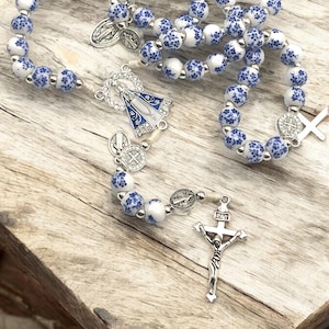 Puede incluir: Un rosario con cuentas blancas y azules con motivos florales. Incluye un crucifijo plateado, una pieza central con una figura religiosa y varios medallones plateados. Las cuentas est&aacute;n ensartadas en un cord&oacute;n blanco.