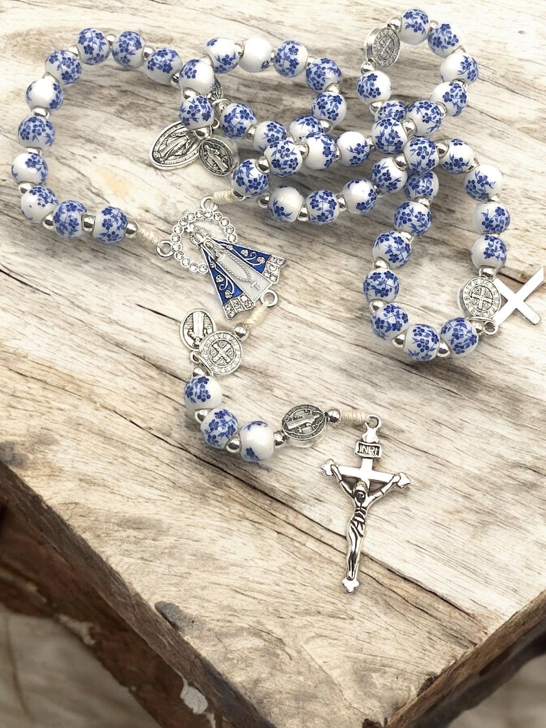 Puede incluir: Un rosario con cuentas blancas y azules con motivos florales. Incluye un crucifijo plateado, una pieza central con una figura religiosa y varios medallones plateados. Las cuentas est&aacute;n ensartadas en un cord&oacute;n blanco.