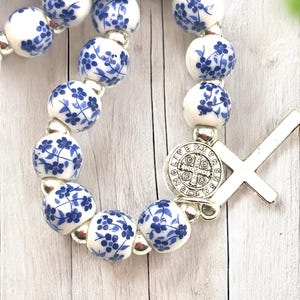 Puede incluir: Un rosario con cuentas de cer&aacute;mica blanca decoradas con motivos florales azules. Las cuentas est&aacute;n unidas con eslabones de metal plateado. Una cruz plateada y un colgante circular con texto est&aacute;n adjuntos.