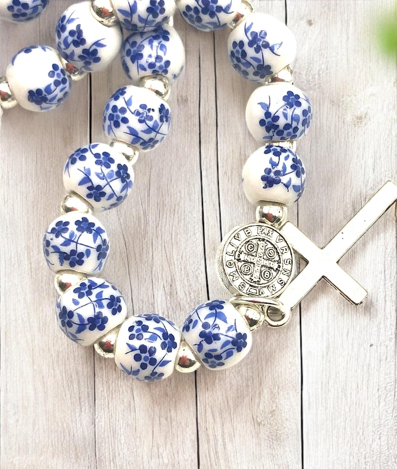 Puede incluir: Un rosario con cuentas de cer&aacute;mica blanca decoradas con motivos florales azules. Las cuentas est&aacute;n unidas con eslabones de metal plateado. Una cruz plateada y un colgante circular con texto est&aacute;n adjuntos.