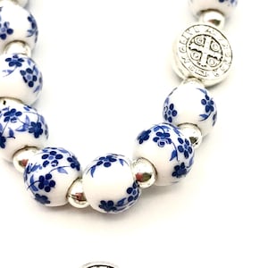 Puede incluir: Primer plano de un rosario con cuentas de porcelana blanca decoradas con motivos florales azules. El rosario presenta detalles met&aacute;licos plateados, incluyendo un colgante en forma de cruz y cuentas de conexi&oacute;n. Las cuentas est&aacute;n ensartadas en un cord&oacute;n blanco.