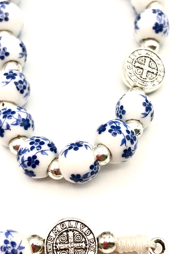 Puede incluir: Primer plano de un rosario con cuentas de porcelana blanca decoradas con motivos florales azules. El rosario presenta detalles met&aacute;licos plateados, incluyendo un colgante en forma de cruz y cuentas de conexi&oacute;n. Las cuentas est&aacute;n ensartadas en un cord&oacute;n blanco.