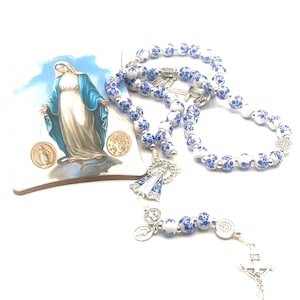Puede incluir: Un rosario con cuentas blancas y azules con estampado floral, un crucifijo plateado y un peque&ntilde;o colgante. Tambi&eacute;n hay una baldosa decorativa con una imagen de la Virgen Mar&iacute;a. El rosario es un art&iacute;culo religioso.