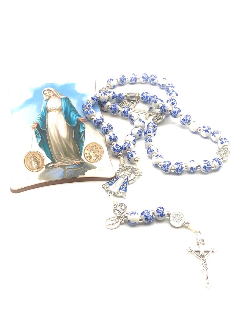 Puede incluir: Un rosario con cuentas blancas y azules con estampado floral, un crucifijo plateado y un peque&ntilde;o colgante. Tambi&eacute;n hay una baldosa decorativa con una imagen de la Virgen Mar&iacute;a. El rosario es un art&iacute;culo religioso.