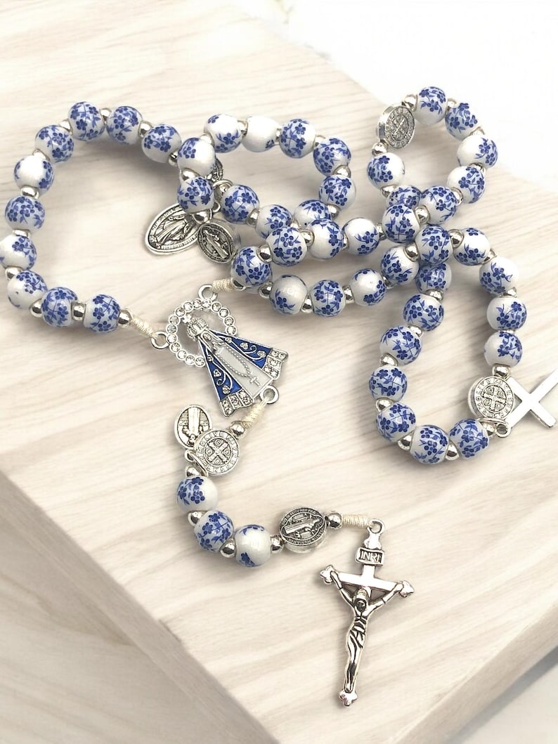 Puede incluir: Un rosario con cuentas blancas y azules con motivos florales. Presenta un crucifijo plateado, una pieza central con una figura religiosa y varios medallones plateados. Las cuentas est&aacute;n unidas por un cord&oacute;n blanco.