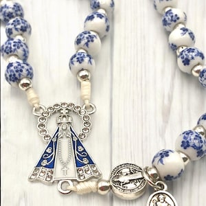Puede incluir: Un rosario con cuentas blancas y azules con motivos florales. El rosario presenta una pieza central plateada con una imagen azul y blanca de la Virgen Mar&iacute;a y una cruz plateada. Las cuentas est&aacute;n unidas por un cord&oacute;n color crema.