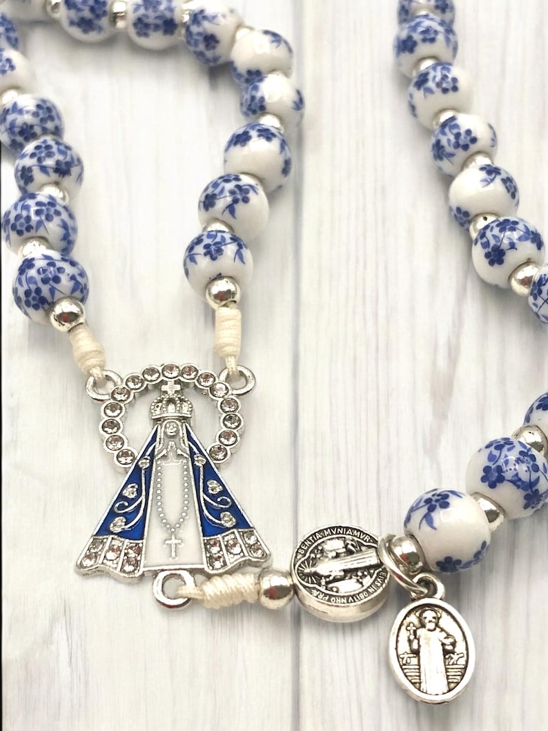 Puede incluir: Un rosario con cuentas blancas y azules con motivos florales. El rosario presenta una pieza central plateada con una imagen azul y blanca de la Virgen Mar&iacute;a y una cruz plateada. Las cuentas est&aacute;n unidas por un cord&oacute;n color crema.