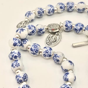 Puede incluir: Un rosario con cuentas blancas adornadas con motivos florales azules. Las cuentas est&aacute;n separadas por espaciadores plateados. Un colgante plateado con una imagen religiosa est&aacute; adjunto.