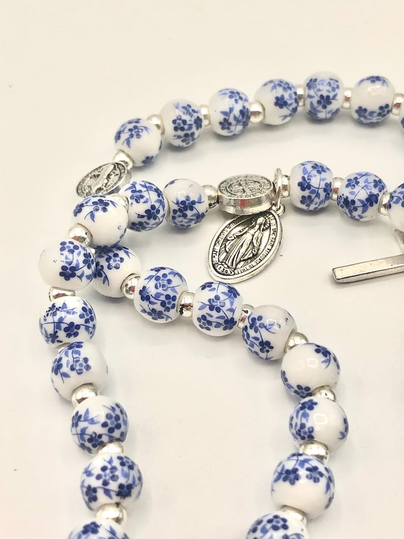 Puede incluir: Un rosario con cuentas blancas adornadas con motivos florales azules. Las cuentas est&aacute;n separadas por espaciadores plateados. Un colgante plateado con una imagen religiosa est&aacute; adjunto.
