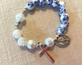 Puede incluir: Pulsera de cuentas con cuentas blancas en forma de rosa y cuentas con estampado floral azul y blanco. Incluye un dije de cruz plateada y una medalla religiosa. Las cuentas están ensartadas en una cadena plateada.
