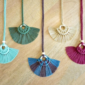 Puede incluir: Cinco collares de macramé con borlas en forma de abanico en diferentes colores, incluyendo turquesa, verde, beige, burdeos y azul y morado. Cada collar presenta una cuenta de madera blanca y un diseño circular de macramé.
