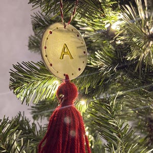 Puede incluir: Adorno navideño circular dorado con la letra "A" en el centro. El adorno tiene una borla roja y cuelga de una cuerda roja y blanca. El adorno está en un árbol de Navidad.