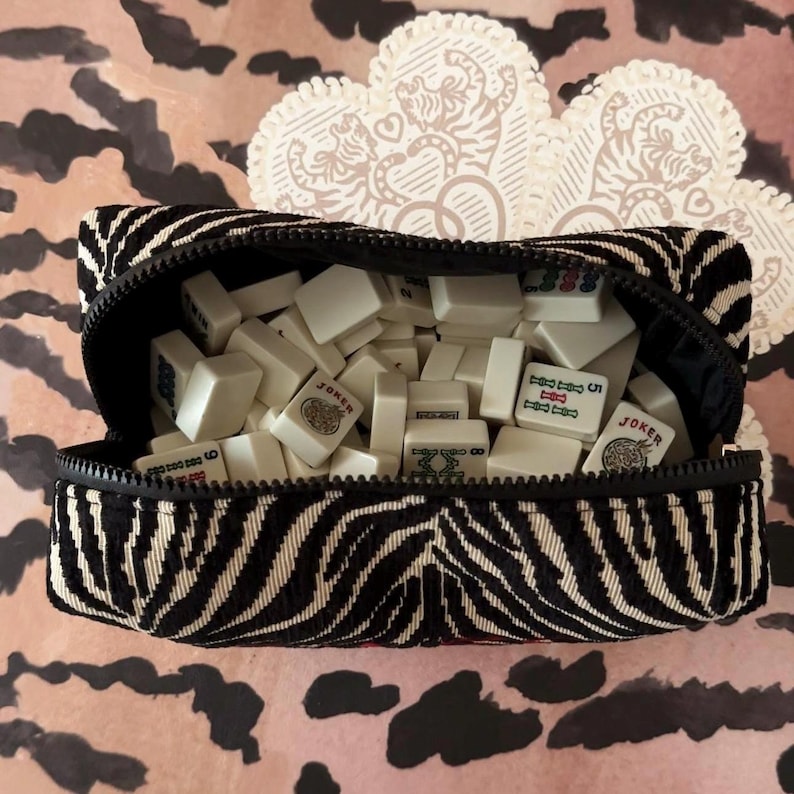 May include: A black and white zebra-striped zippered pouch filled with white Mahjong tiles. The pouch is open, revealing the tiles inside. The background features a pink and brown animal print.