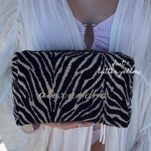May include: A rectangular zippered pouch featuring a black and beige zebra stripe pattern. The name alexandra is embroidered in cursive script on the front. Text on the right side reads font 7 butter yellow. The pouch has a gold-toned zipper pull.