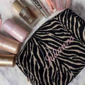 May include: A black and beige zebra-striped cosmetic bag with the name Julianna embroidered in pink script. Surrounding the bag are several skincare and beauty products, including three rhode bottles, a Merit serum, and various lip gloss tubes.