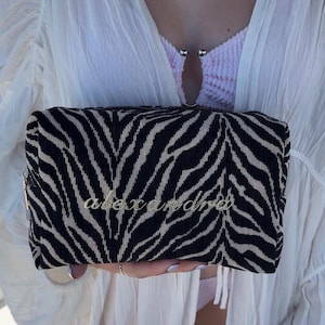 May include: A rectangular zippered pouch featuring a black and beige zebra stripe pattern. The name alexandra is embroidered in a cursive gold script across the center of the front panel. The pouch has a textured fabric surface and a gold zipper pull.