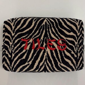 May include: A rectangular cosmetic bag with a zebra stripe pattern in black and beige. The word "TILES" is embroidered in red across the center of the bag. The bag has a soft, textured appearance.