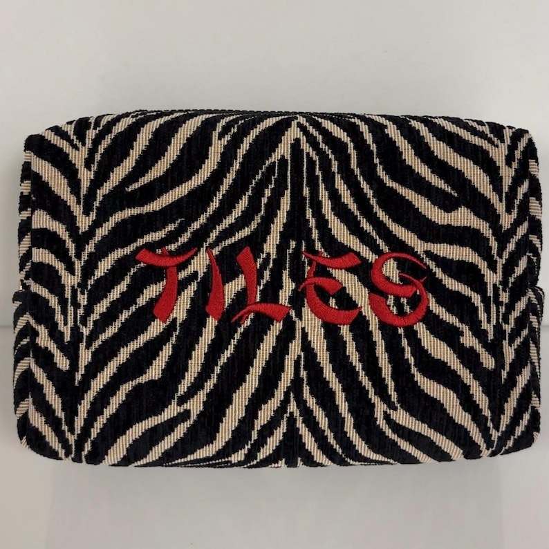 May include: A rectangular cosmetic bag with a zebra stripe pattern in black and beige. The word "TILES" is embroidered in red across the center of the bag. The bag has a soft, textured appearance.