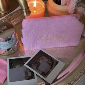 May include: A rectangular light pink zippered pouch with a textured, woven-like surface. The name Alexandra is embroidered in a cursive, light-colored script on the front center of the pouch. The pouch features a matching pink zipper closure.
