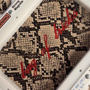 Puede incluir: Un bastidor de bordado blanco con el texto Strong Magnets y U.S. 7,607,399 impreso en los bordes, que sujeta una tela con estampado de piel de serpiente beige y negra con el texto en cursiva roja bordado bag of tricks.
