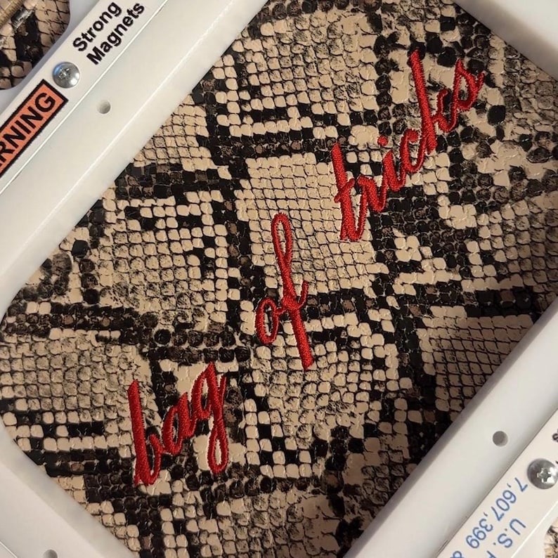 Puede incluir: Un bastidor de bordado blanco con el texto Strong Magnets y U.S. 7,607,399 impreso en los bordes, que sujeta una tela con estampado de piel de serpiente beige y negra con el texto en cursiva roja bordado bag of tricks.