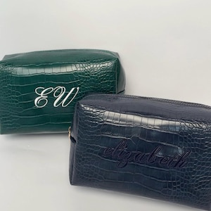 Puede incluir: Dos estuches rectangulares con cremallera y textura de cocodrilo. El estuche verde oscuro presenta las iniciales EW bordadas en blanco, mientras que el estuche azul marino muestra el nombre elizabeth bordado en un hilo oscuro a juego.
