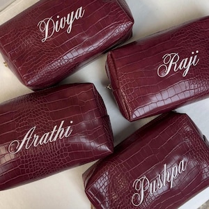 Puede incluir: Cuatro neceseres rectangulares de color burdeos con un patr&oacute;n de textura de piel de cocodrilo. Cada bolsa presenta un nombre bordado en blanco en la parte frontal, concretamente Divya, Raji, Arathi y Pushpa, en letra cursiva.