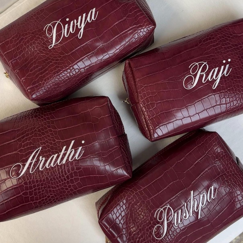 Puede incluir: Cuatro neceseres rectangulares de color burdeos con un patr&oacute;n de textura de piel de cocodrilo. Cada bolsa presenta un nombre bordado en blanco en la parte frontal, concretamente Divya, Raji, Arathi y Pushpa, en letra cursiva.